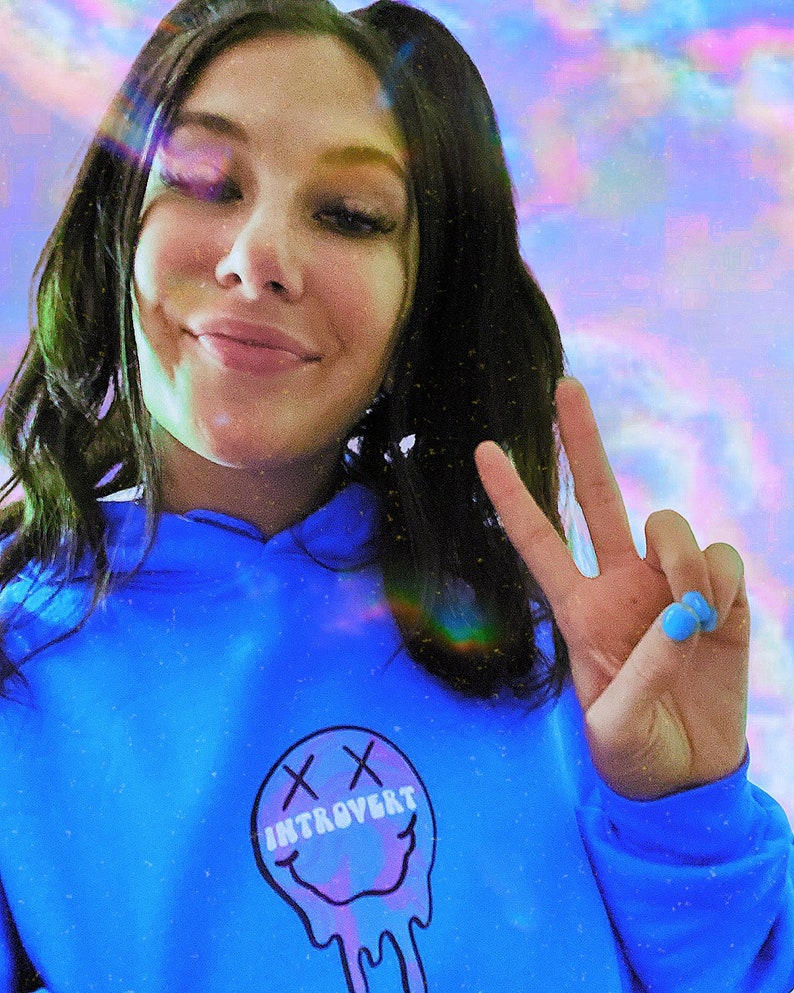 May include: A person wearing a bright blue hoodie with the word "INTROVERT" inside a melting smiley face graphic. The person is giving a peace sign with blue painted nails. The background has a colorful, dreamy effect.