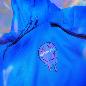 May include: Bright blue hoodie with a drawstring hood and a front graphic. The graphic is a melting smiley face with the word "INTROVERT" in white text. The smiley face has pink and purple accents.