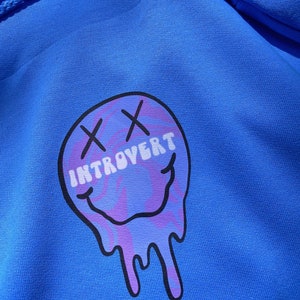 May include: A royal blue hoodie featuring a graphic of a melting smiley face with the word "INTROVERT" in white text. The smiley face has purple and blue swirls and black "X" eyes.