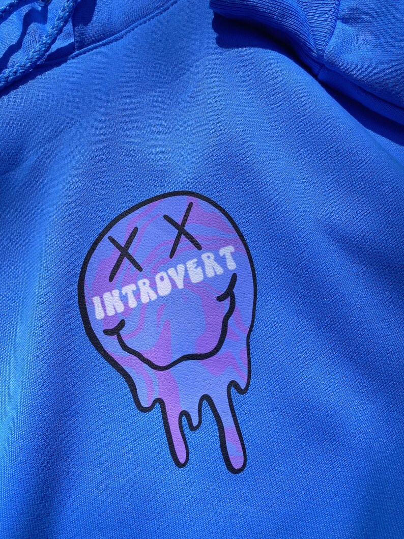 May include: A royal blue hoodie featuring a graphic of a melting smiley face with the word "INTROVERT" in white text. The smiley face has purple and blue swirls and black "X" eyes.