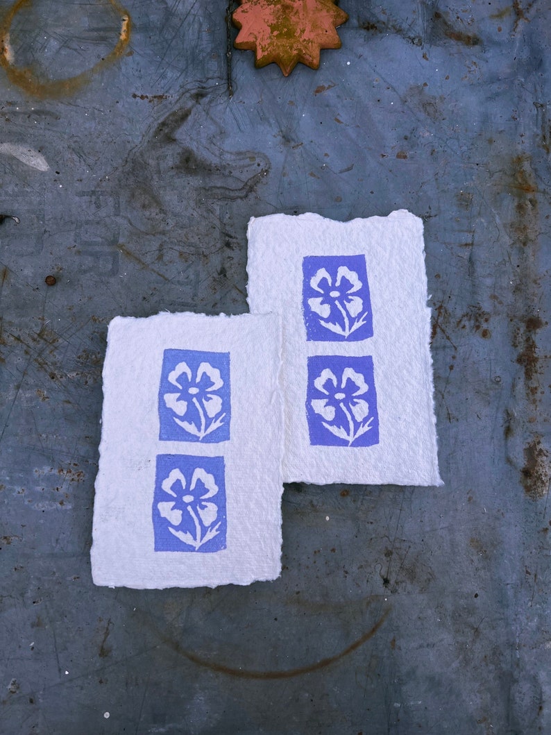 Puede incluir: Dos tarjetas de papel blanco con bordes azules y dise&ntilde;os florales blancos impresos en ellas. Las tarjetas est&aacute;n sobre una superficie de metal gris.