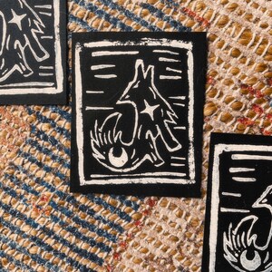 Peut inclure: Estampes linogravées en noir et blanc représentant un renard stylisé. Le renard est représenté avec une étoile et un œil, sur fond noir avec une bordure blanche. Les estampes sont présentées sur une surface texturée.