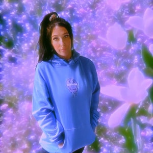 May include: A light blue hoodie with a graphic of a melting smiley face and the word "SLEEPLESS" printed on the front. The hoodie has a kangaroo pocket and a drawstring hood. The background is a blurred floral design in shades of purple and white.
