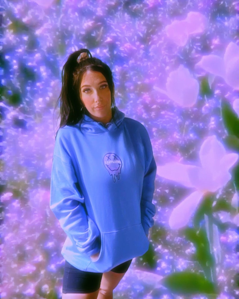 May include: A light blue hoodie with a graphic of a melting smiley face and the word "SLEEPLESS" printed on the front. The hoodie has a kangaroo pocket and a drawstring hood. The background is a blurred floral design in shades of purple and white.