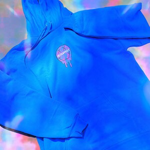 May include: Bright blue hoodie with a front pocket and drawstring hood. The hoodie features a graphic of a melting smiley face with the word "INTROVERT" in a pink and purple color scheme.