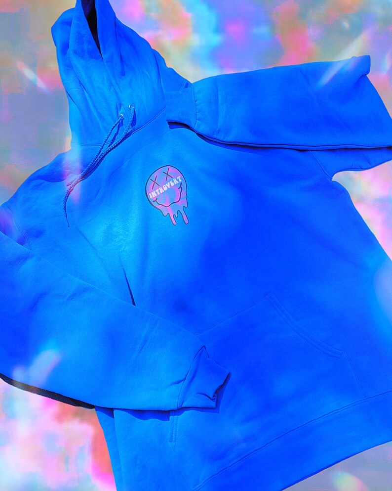 May include: Bright blue hoodie with a front pocket and drawstring hood. The hoodie features a graphic of a melting smiley face with the word "INTROVERT" in a pink and purple color scheme.