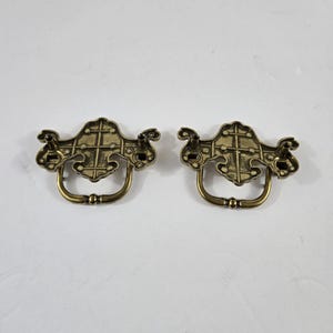 May include: Two antique brass-toned metal drawer pulls featuring a decorative backplate with a grid-like texture and a swinging bail handle. The backplates have a symmetrical, ornate shape with two mounting points on the reverse side.