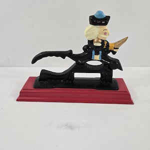May include: A cast iron nutcracker shaped like a figure wearing a black hat with a blue top, a black coat, and white hair. The figure is mounted on a rectangular red base. The nutcracker mechanism is black with a serrated jaw for cracking nuts.