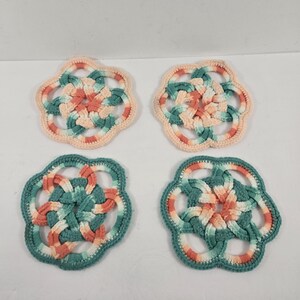 May include: Four handmade crochet coasters. Each coaster features a floral design with interwoven strands of peach, teal, and cream-colored yarn. The coasters are arranged in a square formation against a white background.