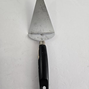 May include: A stainless steel pie server with a triangular blade and a black handle. The handle has a hole at the end for hanging. The server is designed for cutting and serving pies, cakes, and other desserts.