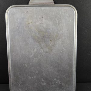 May include: A rectangular, aluminum baking pan with a handle. The pan has rounded corners and a slightly textured surface. The handle is a small, curved piece of metal attached to one end. The pan shows signs of wear and use.