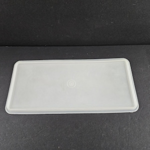 May include: A rectangular, translucent plastic lid with a slightly raised edge. The lid is a light, neutral color and has a small, circular indentation in the center. The lid is designed to fit a rectangular container.