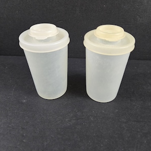 May include: Two translucent plastic containers with off-white lids. The containers are cylindrical with a slightly tapered shape. The lids are circular and fit snugly on top. The background is a solid black.