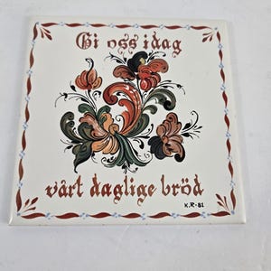 May include: Square ceramic tile with a floral design in shades of orange, green, and brown. The tile has text in a script font that reads "Gi oss idag" and "vårt daglige bröd". The border is decorated with small blue and brown accents.
