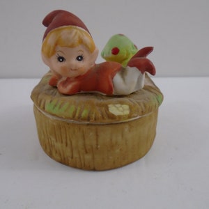 May include: A ceramic trinket box featuring a whimsical elf figure in a red outfit and hat, lying on a straw-like base. The box has a light brown, textured exterior and a small green and red accent.