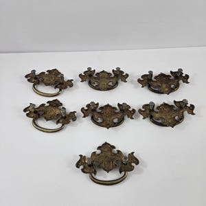 May include: Seven brass-toned metal drawer pulls with ornate, decorative backplates and swinging bail handles. Each pull features a symmetrical, detailed design with two mounting screws attached to the backplate for secure furniture installation.