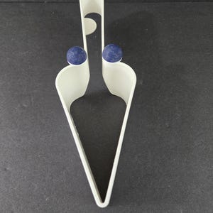 May include: A white, plastic cake server with a unique design. The server features a handle with a curved top and two blue spheres. The serving end is a pointed triangle shape. The server is on a dark surface.