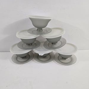May include: A set of six translucent gray plastic dessert bowls with matching white lids, arranged in a pyramid stack. Each bowl features a wide circular base, a short pedestal stem, and a rounded basin, with the Tupperware brand name visible.