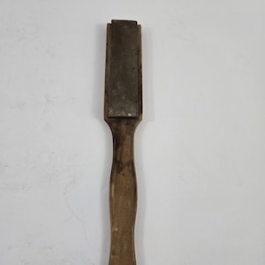 May include: A vintage sharpening stone with a wooden handle. The rectangular stone is set in a wooden frame and is a dark gray color. The handle is a light brown color and has a hole at the end.