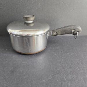 May include: A small, stainless steel saucepan with a lid and a black handle. The pot has a rounded base and a lid with a small, round, black knob. The handle is attached with rivets and has a metal loop at the end.