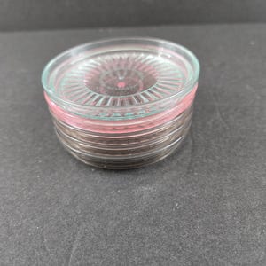 May include: A stack of clear glass coasters with a pink rim and a textured, radial design. The coasters are circular and appear to be made of glass, with a pink accent on the edge.