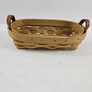 May include: A light brown, oval-shaped basket with a woven design. The basket has two brown leather handles and is made of wood. The basket is empty and sits on a white surface.