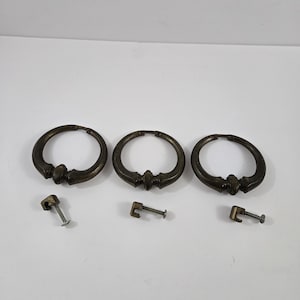 May include: Three circular brass drawer pulls with a dark, aged patina finish. Each pull features a central decorative detail at the bottom and is accompanied by a matching metal mounting bracket and screw for installation on furniture pieces.