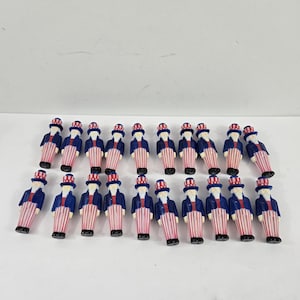 May include: A collection of small Uncle Sam figurines. Each figurine is dressed in a blue coat, red and white striped pants, and a top hat. The figurines are arranged on a white surface.