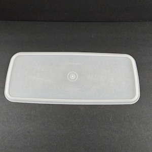 May include: A clear, rectangular Tupperware lid. The lid is made of a translucent plastic material and has a slightly raised edge. The Tupperware logo is visible in the center of the lid.