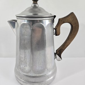 May include: A vintage aluminum stovetop coffee maker with a wooden handle and lid knob. The coffee pot has a classic design with a spout and a rounded base. The handle is a dark brown color, and the lid knob is also made of wood.