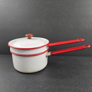 May include: A white enamel double boiler set featuring a red rim and two long red handles. The top pot includes a matching white lid with a small brown knob. The cookware has a smooth finish with visible red accents along the edges and handles.