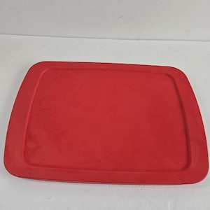 May include: A rectangular, red Pyrex lid. The lid is a solid, vibrant red color with a slightly curved edge. The Pyrex logo is visible in the upper left corner. The lid is designed to fit a rectangular Pyrex dish.