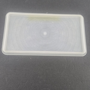 May include: A clear, rectangular Tupperware lid with a white rim. The lid has a slightly translucent appearance and features a circular design in the center. The Tupperware logo is visible. The lid is designed to fit a rectangular container.