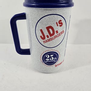 May include: A white and gray travel mug with a blue handle, lid, and accents. The mug features the text "J.D.'s Hamburgers" in a red circle, along with a 25th-anniversary logo. The mug has a textured surface.