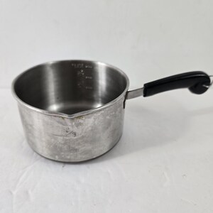 May include: A small, stainless steel saucepan with a black handle. The pan has measurement markings inside and shows signs of wear. The handle is curved and attached to the side of the pan.