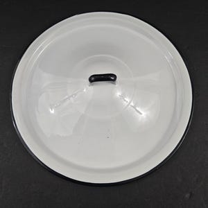 May include: A white enamel lid with a black rim and a small, black handle. The lid is round and reflects light, suggesting a glossy finish. The item appears to be a kitchenware accessory.