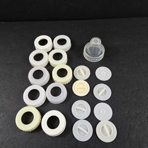 May include: An assortment of off-white and clear plastic bottle parts, including screw-on rings, round caps, and a nipple. The parts are arranged on a black surface, suggesting a collection of baby bottle components.