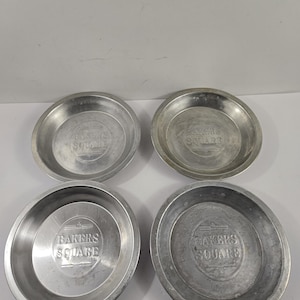 May include: Four round, silver-colored metal pie pans. Each pan has a slightly raised rim and the words "BAKERS SQUARE" embossed in the center. The pans are arranged in a square formation against a white background.