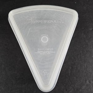 May include: A clear, triangular Tupperware container lid. The lid has a raised edge and the Tupperware logo is visible. The text "Made in U.S.A." is also present. The lid is made of a translucent plastic material.