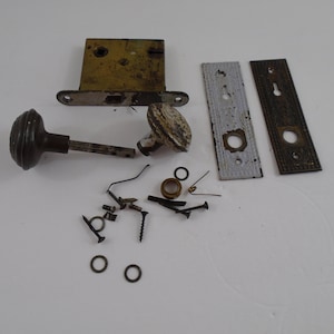 May include: A vintage door hardware set with a brass door plate, a white porcelain door knob, a black door knob, and a brass door lock. The set includes screws, springs, and other hardware.