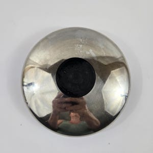 May include: A circular, polished silver-tone metal lid featuring a smooth, reflective surface and a centered, circular black knob. The metallic finish displays a high-gloss sheen, and the design is simple, functional, and consistent in its round shape.