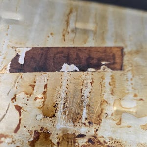 May include: Close-up of a stained, off-white surface with brown discoloration and a rectangular, brown label. The surface shows signs of wear and tear, with scratches and peeling. The overall appearance suggests age and use.