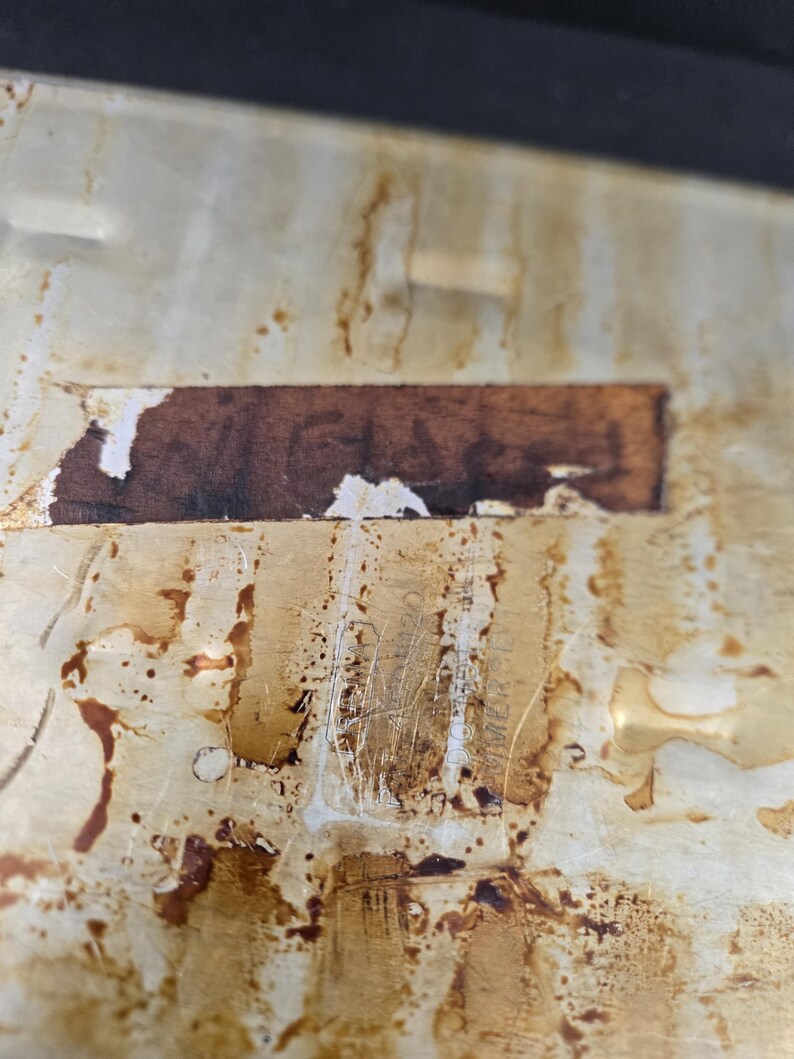 May include: Close-up of a stained, off-white surface with brown discoloration and a rectangular, brown label. The surface shows signs of wear and tear, with scratches and peeling. The overall appearance suggests age and use.