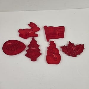 May include: A collection of red plastic cookie cutters in various shapes, including a bunny, an American flag, a snowman, a holly leaf, a Christmas tree, and an egg with the words "Happy Easter".