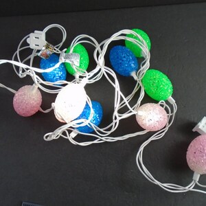 May include: A string of Easter egg-shaped lights in various colors, including pink, blue, green, and white. The lights are connected by a white wire and have a textured, glittery surface. The plug is white.