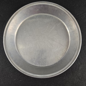May include: A round, silver-colored metal pie pan. The pan has a slightly raised edge and a smooth, reflective surface with visible scratches. The pan is designed for baking and serving pies.