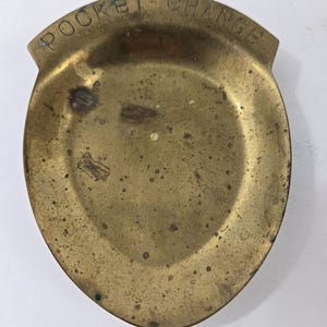 May include: A vintage brass pocket change dish. The dish is shield-shaped with a rounded bottom and a small, raised top. The words "POCKET CHANGE" are embossed across the top. The brass has a tarnished, aged appearance.