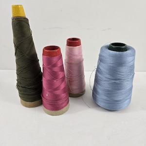 May include: Four spools of thread in various colors. One is dark green with a yellow thimble, two are pink, and one is light blue. The spools are on cardboard bases, ideal for sewing and crafting projects.
