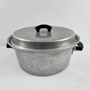 May include: A vintage aluminum cooking pot with a lid and black handles. The pot is round with a slightly textured surface and a black handle on the lid. The pot is designed for cooking.