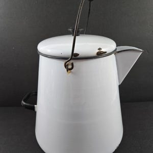 May include: White enamelware coffee pot with a black handle and spout. The pot has a black rim and a wire handle with a wooden grip. A classic kitchenware item, perfect for camping or home use.